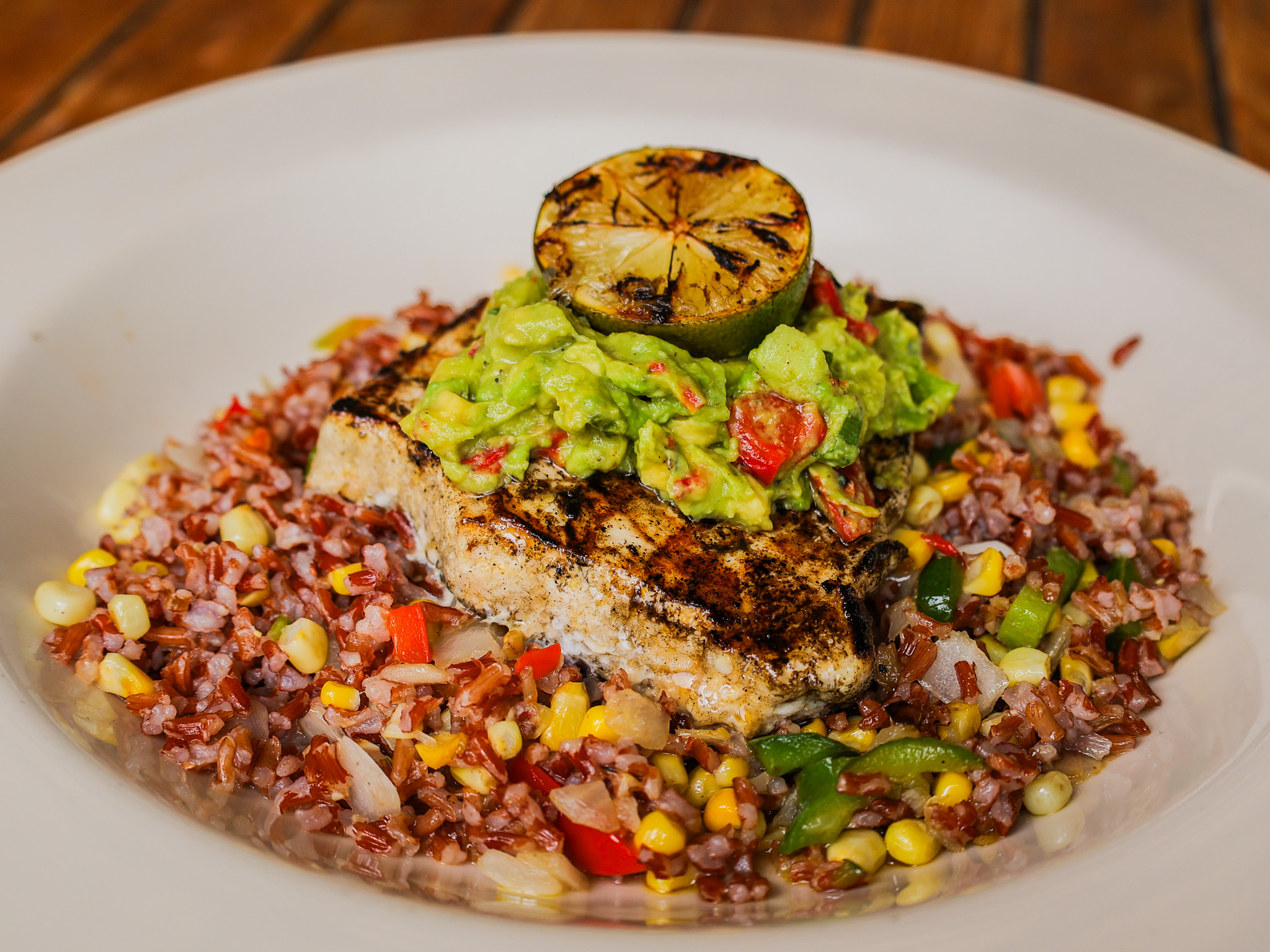 A grilled swordfish with a vibrant green avocado salad and a grilled lime, sitting on top of a bed of red rice tossed with corn.