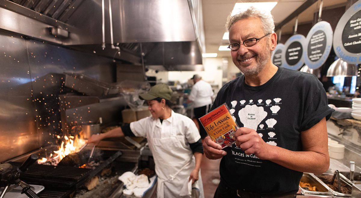 Ari Weinzweig holds up his new chapbook in front of the kitchen at the Roadhouse, where a line cook flames up a pan over the burner.