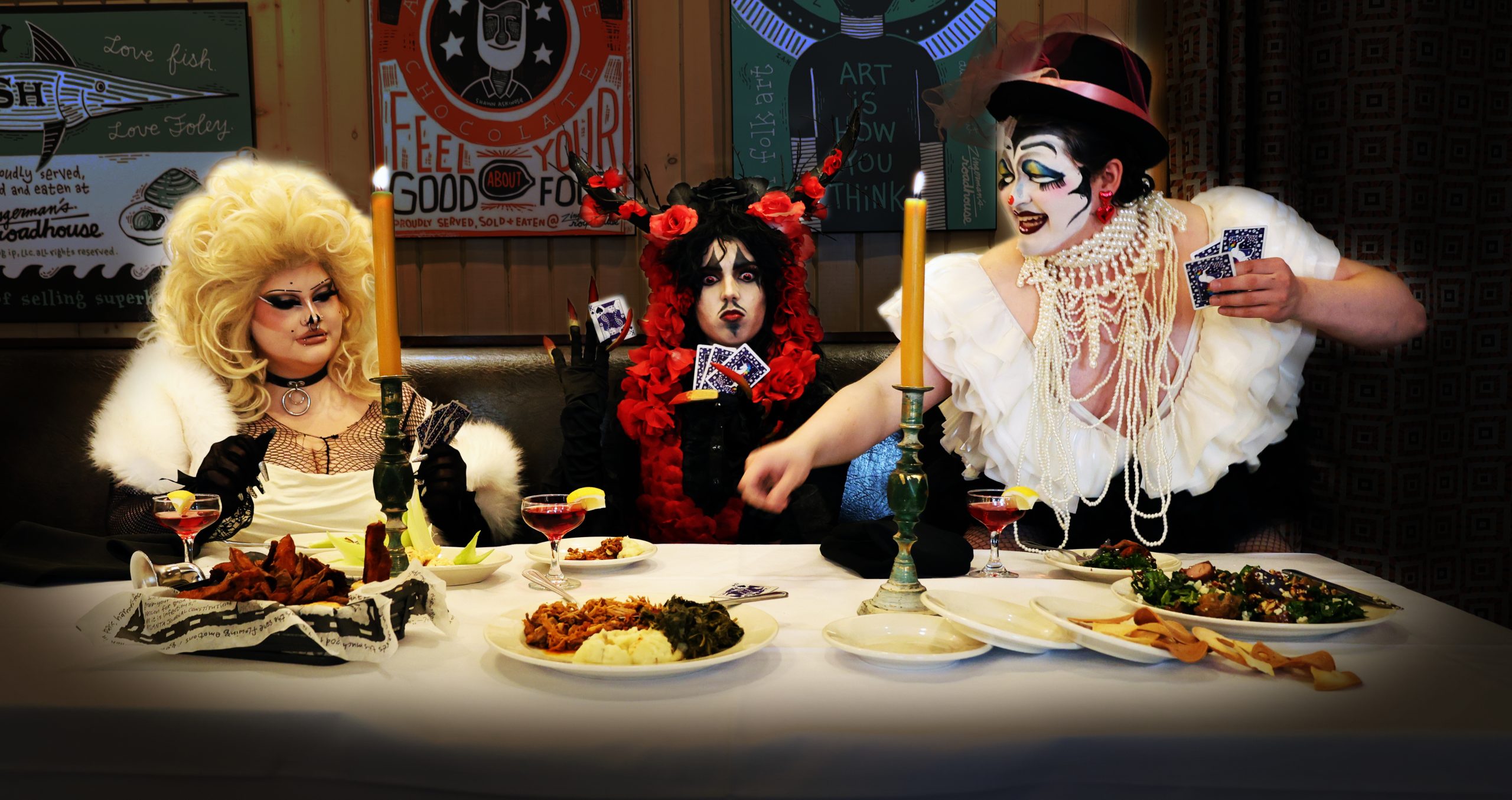 A group of drag queens surrounds a table with food, playing cards